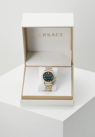 Versace-klocka med grön urtavla, guldfärgad boett och silver-guld metallarmband, presenterad i en vit ask med gyllene insida.