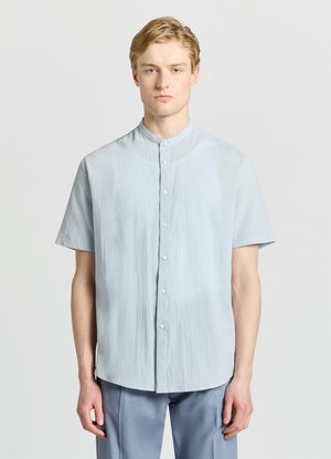 Junger Mann mit kurzen blonden Haaren, der ein hellblaues kurzärmeliges Hemd und blaue Hose trägt, steht vor einem einfarbigen Hintergrund.