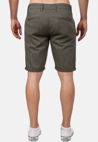 Olijfgroene shorts met opgerolde manchetten, twee achterzakken, contrasterende rode stiksels en een label op de tailleband. G gedragen met witte sneakers.