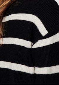 Pull en tricot noir avec de larges bandes horizontales blanches, présentant une texture côtelée et une coupe décontractée, mettant l'accent sur la zone des épaules.