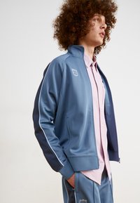 Chaqueta atlética azul con cremallera frontal, rayas laterales y logotipo bordado. La lleva puesta sobre una camisa de botones a rayas rosas, combinada con pantalones a juego.