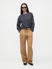 Femme portant des lunettes de soleil noires, un t-shirt à manches longues rayé noir et blanc, un pantalon ample beige avec un cordon et des chaussures noires, debout les mains dans les poches.