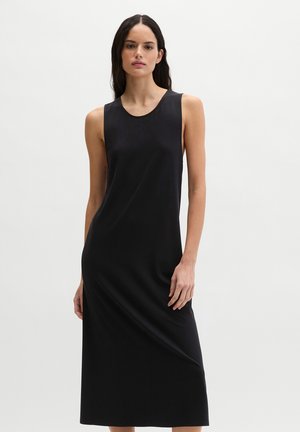 Kobieta o długich ciemnych włosach, ubrana w czarną, bezrękawową sukienkę midi, stojąca na tle jednolitego białego tła.