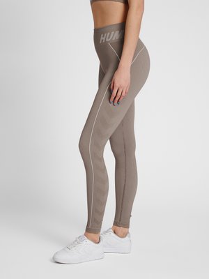 Femme portant des leggings taupe taille haute avec un motif chevron et des baskets blanches, debout de profil sur un fond uni.