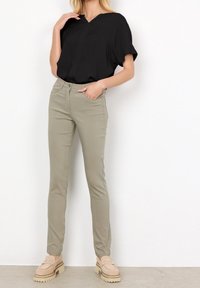 Femme debout les mains dans les poches, portant un blouse noire à manches courtes, un pantalon beige clair slim-fit, et des mocassins à plateformes beiges sur un sol gris.