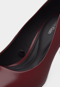 Γυναικείο ψηλοτάκουνο παπούτσι από σκούρο κόκκινο δέρμα με μαύρη επένδυση εσωτερικά και επωνυμία Calvin Klein.
