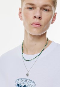 Witte T-shirt met blauwe geborduurde letters, gelaagd met twee halskettingen: één zilveren ketting met een hanger en één meerkleurige kraalstrang.