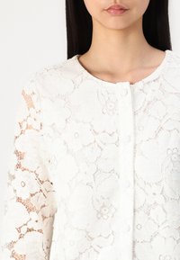 Blusa bianca in pizzo con motivi floreali, scollatura tonda e chiusura con bottoni. Maniche lunghe con una texture in pizzo intricato.