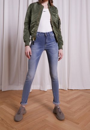 Osoba w zielonej kurtce typu bomber, białym topie Nike, jasnoniebieskich obcisłych dżinsach z podwinięciami i szarych trampkach stoi na drewnianej podłodze.