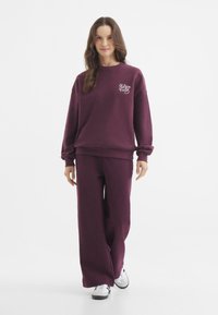 Bordeaux sweatshirt en bijpassende wijde broek van zachte stof, met een klein wit logo op de borst, gedragen met witte sneakers.