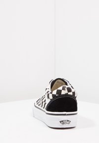 Des chaussures slip-on à carreaux noir et blanc avec des accents en daim, une semelle en caoutchouc blanche, des coutures contrastées et le logo Vans sur le talon.