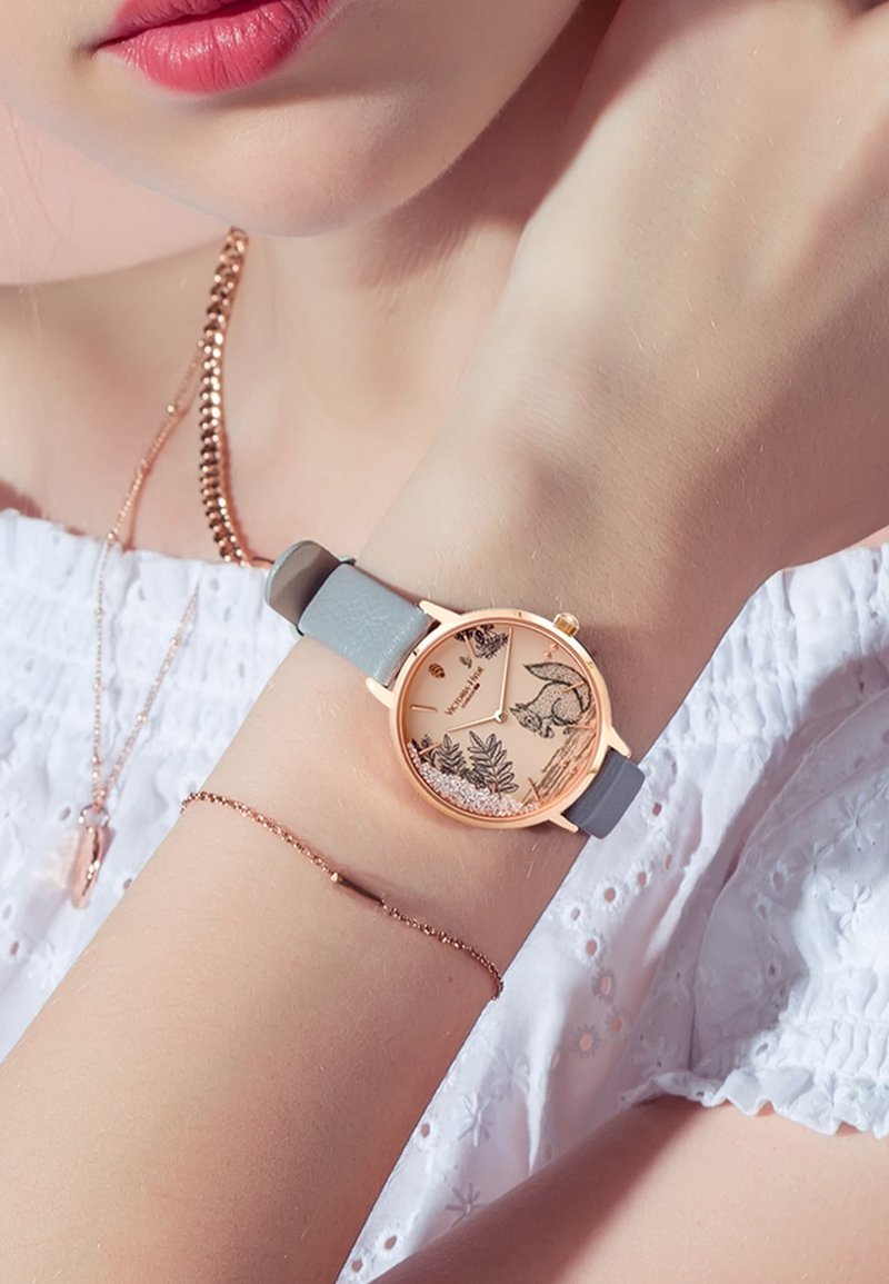 Reloj de oro rosado con correa gris claro; presenta una esfera con un patrón floral y un diseño de zorro; caja redonda; acentos de hardware minimalistas.