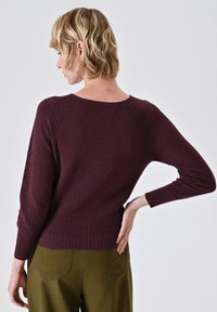 Maglione a maglia bordeaux con scollatura rotonda e polsini a costine, caratterizzato da un design texturizzato nella parte superiore e una cintura aderente, indossato con pantaloni verde oliva.