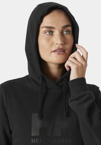 Sudadera negra con capucha ajustable y bolsillo canguro en la parte frontal. Presenta un logo en un tono gris oscuro mate con las letras "HH" y "HELLY HANSEN" impresas en el pecho.