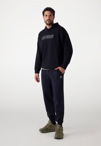 Felpa nera con logo "GUESS", abbinata a pantaloni della tuta scuri con una toppa del logo. Il modello indossa scarpe da ginnastica verde oliva.