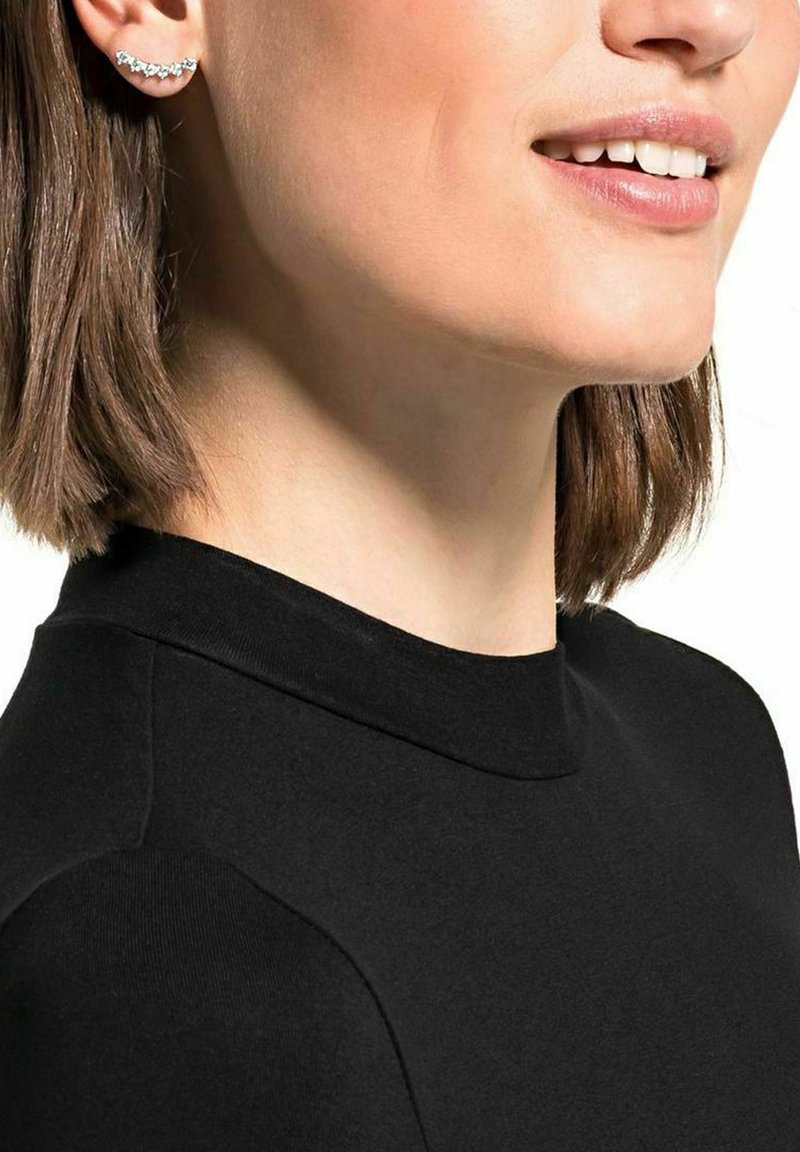 Pendiente de oreja plateado con un diseño curvado y ondulado, adornado con pequeñas piedras transparentes. Se lleva con una blusa negra ajustada. Se ve el cabello corto y liso.