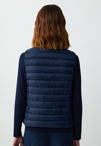 Marineblaue Steppweste mit gestepptem Design und horizontalen Nähten. Leicht, mit glatter Textur, ohne Ärmel, über einem dunklen Pullover getragen.