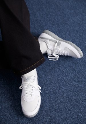Osoba nosząca białe sneakersy marki Nike z szarymi podeszwami i białymi sznurowadłami, czarne spodnie i białe skarpetki na niebieskim wyłożonym dywanem podłodze.