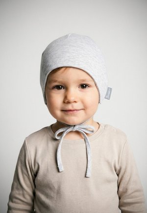 Toddler wearing a light grey bonnet tied under the chin and a beige long-sleeve shirt, facing forward with a neutral background.
