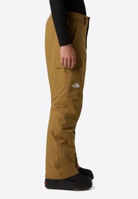 Brune vandtætte udendørsbukser med cargo-lommer og et tætsiddende design, der har et lille hvidt logo på låret og en sort accent ved kanten.