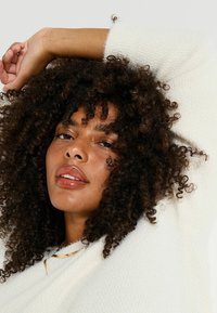 Suéter blanco texturizado con un escote amplio. La modelo tiene el cabello rizado y oscuro y lleva un collar sencillo de oro. Su piel tiene un brillo natural.