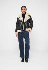 Schwarze Leder-Cropped-Jacke mit cremefarbenem Shearling-Kragen und -Ärmeln, mit Reißverschluss, getragen über einem schwarzen Top und weiten blauen Jeans-Cargo-Hosen.