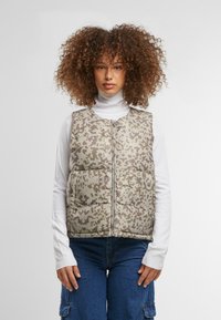 Gepolsterte Weste in einem gedämpften Khaki- und Braun-Tarnmuster, mit Reißverschluss, hohem Kragen und ausgestelltem Saum. Getragen über einem weißen Rollkragenpullover.