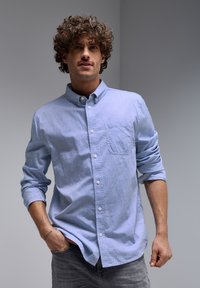 Light blue button-up shirt with a chest pocket, long sleeves rolled up, and a textured fabric. Paired with gray jeans.