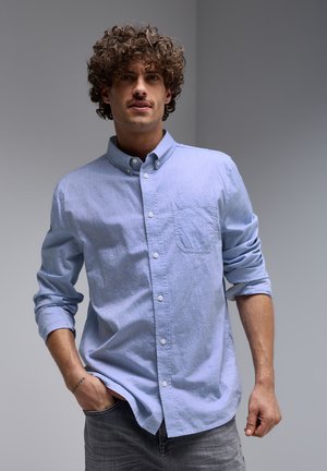 Camicia azzurra chiara con taschino sul petto, maniche lunghe arrotolate e tessuto strutturato. Abbinata a jeans grigi.