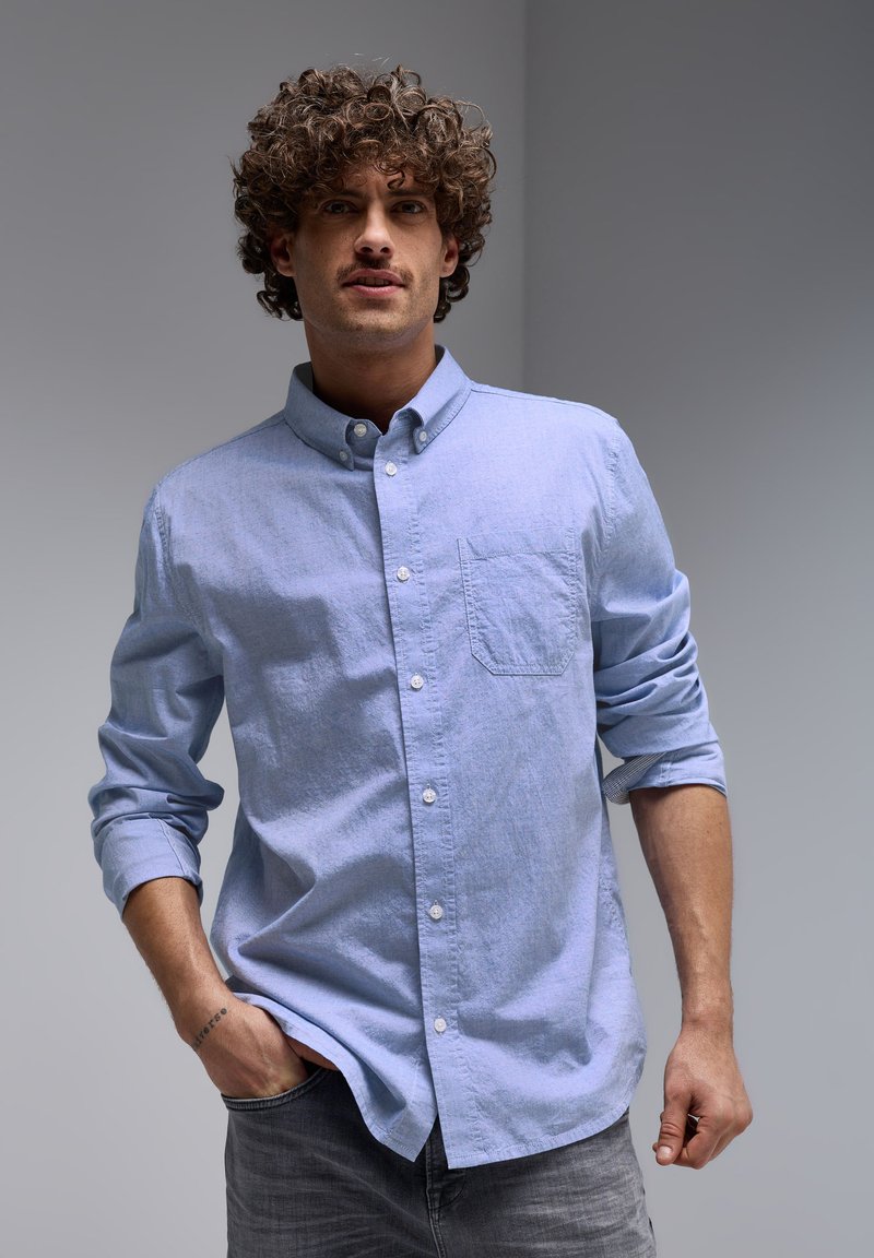Light blue button-up shirt with a chest pocket, long sleeves rolled up, and a textured fabric. Paired with gray jeans.