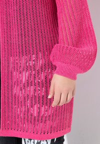 Cardigan rosa lavorato a maglia caratterizzato da un design ampio e aperto e da polsini a coste. Il tessuto ha una texture leggermente lucida ed è abbinato a un completo scuro.