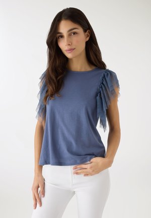 Donna con lunghi capelli scuri che indossa una maglia blu con maniche a volant stratificati in tulle e pantaloni bianchi, in piedi davanti a uno sfondo bianco semplice.