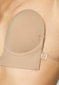 Soutien-gorge adhésif beige avec des bonnets moulés et une texture lisse. Dispose d'attaches en plastique transparent pour un maintien sécurisé. Détails de couture visibles le long des bords.