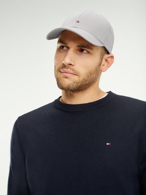 Young man with short beard wearing light gray cap and navy sweater with small red, white, and blue logo on chest, looking upward.