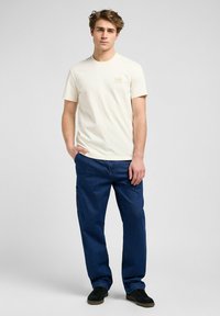 Cremefarvet bomulds t-shirt med kortærmer, prydet med et lille logo på brystet, parret med løstsiddende mørkeblå jeans og sorte sko.