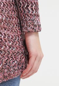 Ruka odpočívající vedle boků, oblečená v růžovém, hnědém a bílém silně pleteném svetru s žebrovanými manžetami a modrých džínách na bílém pozadí.