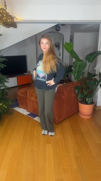 Zwarte sweatshirt met grafisch ontwerp, olijfgroene broek en witte sneakers. Op de achtergrond zijn een plant, een bank en een televisie te zien.