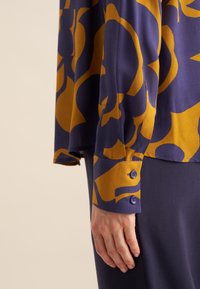 Lange mouwen blouse in paars met gedurfde gele bloemenpatronen. Heeft een knoopmanchet met twee knopen en een relaxte pasvorm.