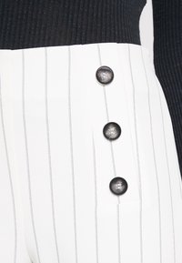 Valkoiset raidalliset housut, joissa on korkea vyötärö, ja neljä suurta mustaa nappia sivussa, sekä teksturoitu musta ribbipaita.