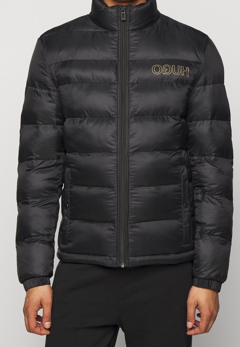 Giacca a vento nera con colletto alto, design trapuntato, zip frontale e dettaglio del logo in oro. Presenta polsini elasticizzati e tasche laterali.