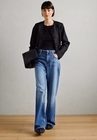 Veste noire texturée, haut noir ajusté, jean bleu taille haute, coupe évasée, et chaussures noires, sur fond neutre.