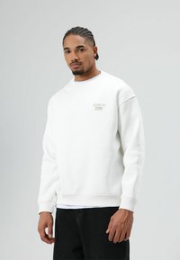Witte sweatshirt gemaakt van katoenmix, met een ronde hals, geribbelde manchetten en een klein geborduurd logo op de borst. Zwarte broek eronder gedragen.