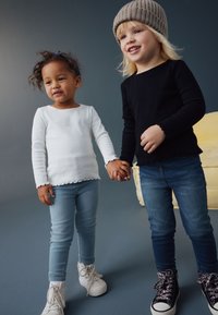 To børn holder hinanden i hånden. Den ene har en hvid ribbet langærmet top og lyseblå jeans, og den anden har en sort langærmet top og mørke jeans.