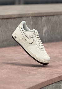 Nike Air Force 1 športni copati iz off-white usnja, s črnimi detajli, perforiranim prstnim delom, ravnimi vezalkami in teksturiranim gumijastim podplatom.