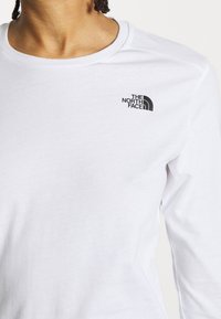 Biała koszula z długim rękawem o okrągłym dekolcie, z małym czarnym logo "The North Face" na górnej lewej części klatki piersiowej. Miękka w dotyku.