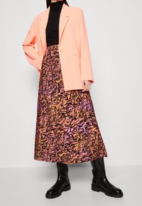 Tenue superposée avec un blazer oversize corail et un col roulé noir, associé à une longue jupe à motifs aux teintes orange, rose et noir. Bottes noires.