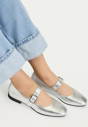 Mujer con zapatos Mary Jane plateados con hebilla, combinados con vaqueros azules claros con puños, sobre un fondo blanco.
