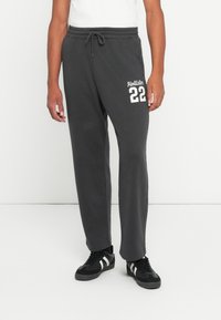 Muškarac nosi tamno siva trenerica s natpisom "Hollister 22" na bedru, uparena s crnim tenisicama s bijelim prugama.