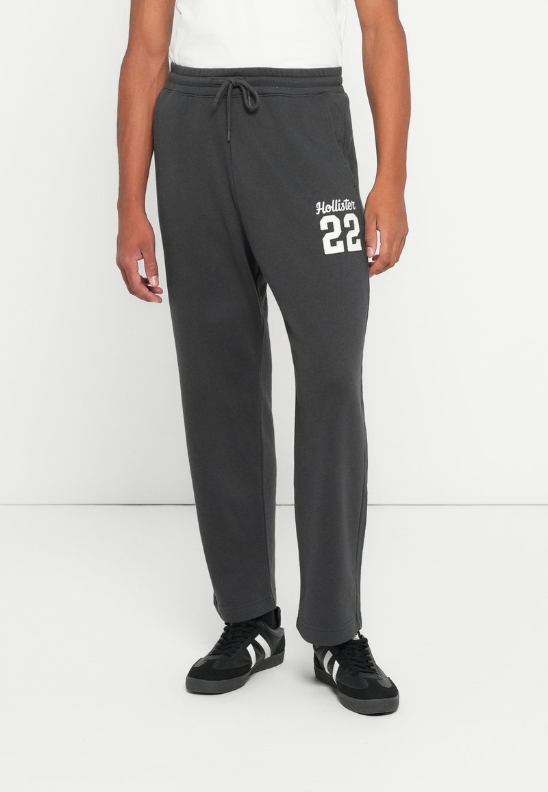 Muškarac nosi tamno siva trenerica s natpisom "Hollister 22" na bedru, uparena s crnim tenisicama s bijelim prugama.