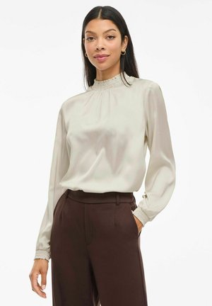 Woman with long dark hair wears a cream high-neck blouse with smocked cuffs and dark brown high-waisted pants, posing with one hand in pocket.